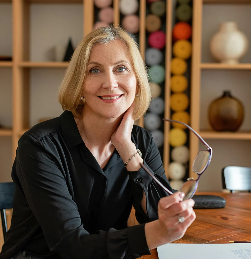 A professional blonde woman wearing glasses and a black blouse sitting at a wooden conference table, smiling warmly next to a clipboard and a modern bookshelf.